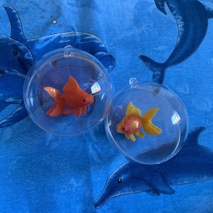 Puede incluir: Dos bolas de pl&aacute;stico transparente, cada una con un peque&ntilde;o pez dorado naranja y amarillo. Las bolas tienen un lazo para colgar y est&aacute;n colocadas sobre un fondo azul con dise&ntilde;os de delfines.