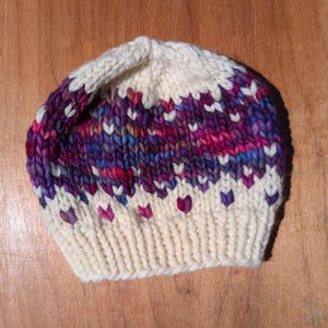 May include: A hand-knitted beanie hat in cream, with a pattern of purple, pink, and blue hearts and stripes. The hat is made of thick yarn and has a ribbed brim. The hat is a winter accessory.
