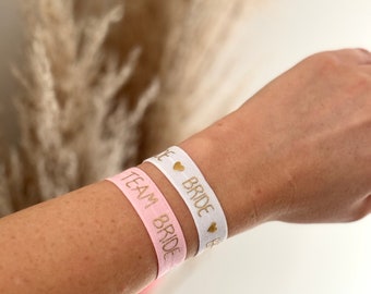 May include: Two elastic wristbands, one pink and one white, are worn on a wrist. The pink band reads "TEAM BRIDE" in gold lettering. The white band says "BRIDE" with gold heart accents. The background features dried ornamental grass.