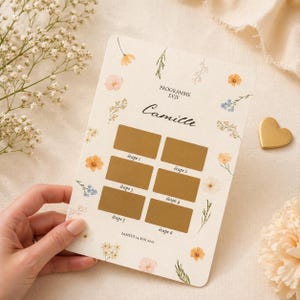 May include: A cream-colored card with floral accents and the text "PROGRAMME EVJF Camille". It features six scratch-off boxes labeled "étape 1" through "étape 6". The date "SAMEDI 24 MAI 2023" is printed at the bottom. A gold heart-shaped charm is visible.