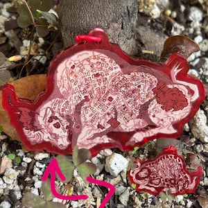 Könnte beinhalten: Ein roter, kartenförmiger Schlüsselanhänger mit einer passenden roten Schlaufe. Der Schlüsselanhänger zeigt ein detailliertes Kartendesign in Rot- und Weißtönen. Eine kleinere Version des Schlüsselanhängers ist ebenfalls sichtbar.