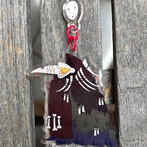 May include: A clear acrylic keychain featuring a stylized illustration of a figure in a dark cloak with white bone-like details. The keychain has a red clasp and a clear top piece with two small holes. The figure's cloak is a gradient of dark red, gray, and blue.