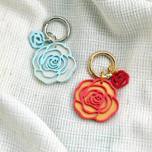 May include: Two keychains featuring layered rose-shaped charms. One keychain has a silver-tone ring with a light blue and white rose charm and a small light blue rose charm. The other has a gold-tone ring with a red and orange rose charm and a small red rose charm.