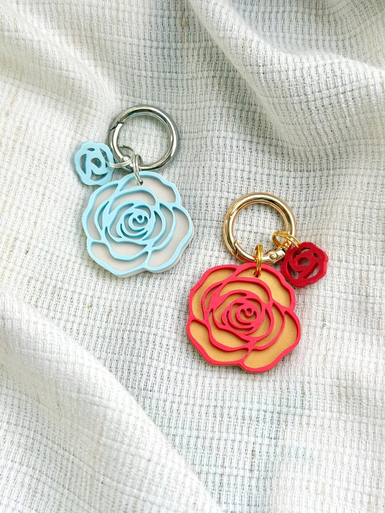May include: Two keychains featuring layered rose-shaped charms. One keychain has a silver-tone ring with a light blue and white rose charm and a small light blue rose charm. The other has a gold-tone ring with a red and orange rose charm and a small red rose charm.