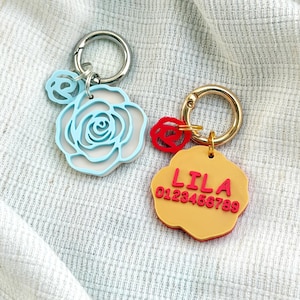 May include: Two keychains with metal rings. One features a light blue rose-shaped charm with a smaller matching rose accent. The other is a yellow flower-shaped charm with a red rose accent, displaying the red text LILA and 0123456789.