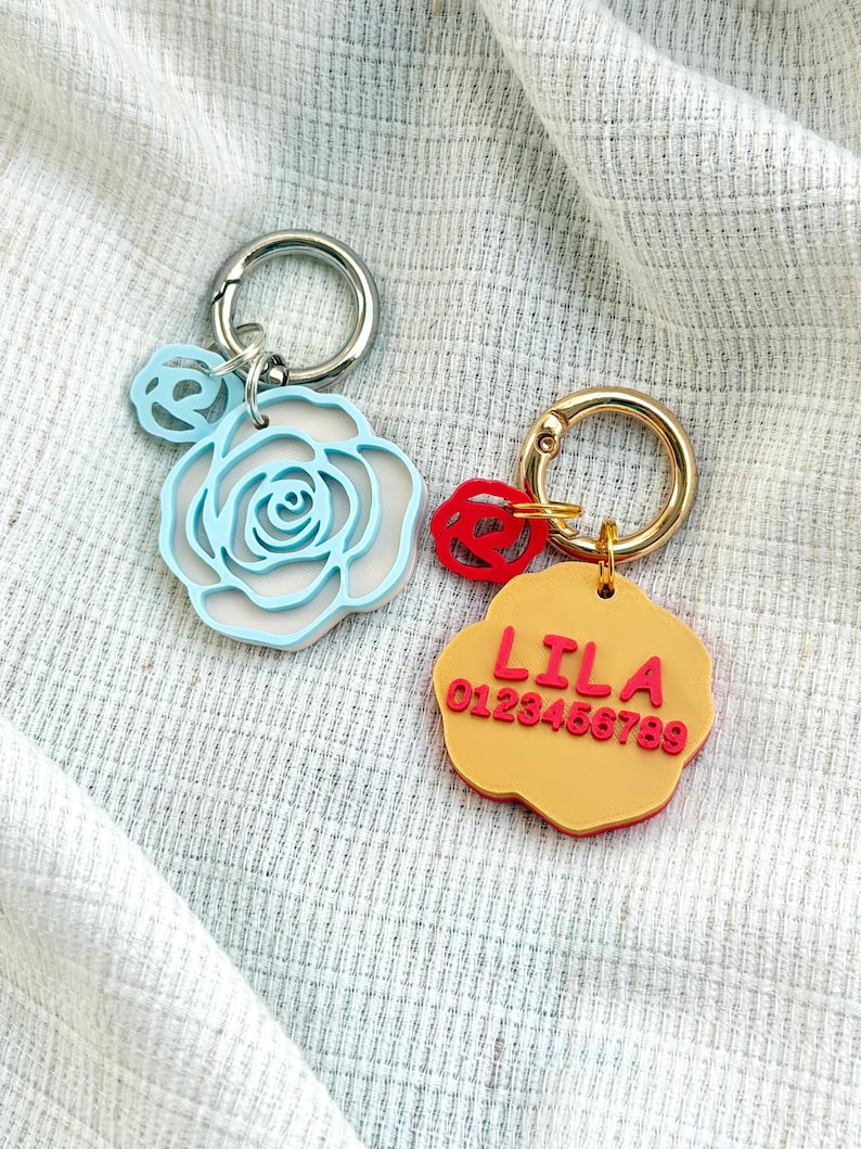 May include: Two keychains with metal rings. One features a light blue rose-shaped charm with a smaller matching rose accent. The other is a yellow flower-shaped charm with a red rose accent, displaying the red text LILA and 0123456789.