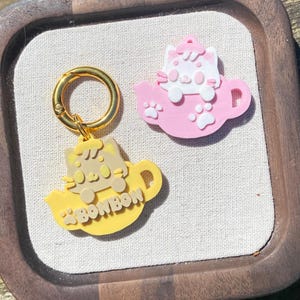 Może przedstawiać: Dwa żółte i różowe breloczki w kształcie kota z złotym metalowym kółkiem. Żółty brelok ma kota siedzącego w czajniku z napisem "Bonbon" z boku. Różowy brelok ma kota siedzącego w czajniku z białymi odciskami łap.