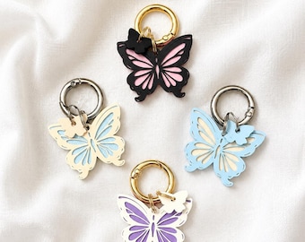 May include: Four butterfly-shaped keychains with metal rings. One is black with pink, one is cream with light blue, one is light blue with cream, and one is white with purple. Each features a smaller butterfly charm attached to the ring.