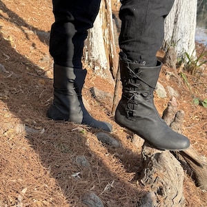 May include: A pair of black leather boots with laces up the front. The boots are worn by a person standing on a tree root in a forest setting.