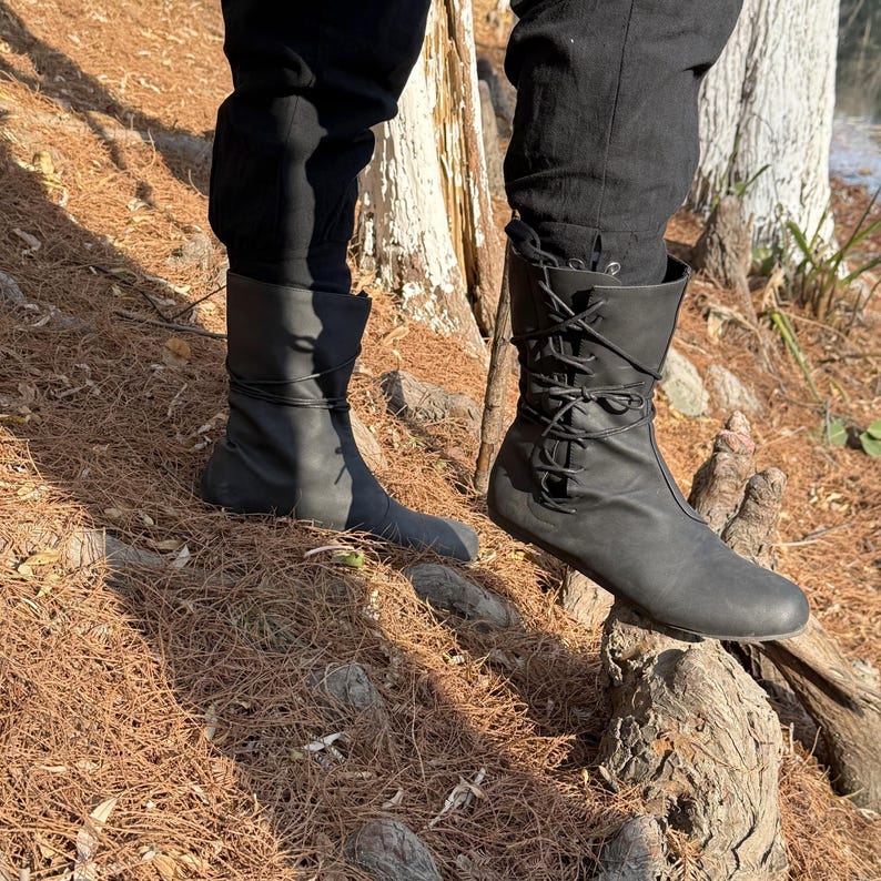May include: A pair of black leather boots with laces up the front. The boots are worn by a person standing on a tree root in a forest setting.
