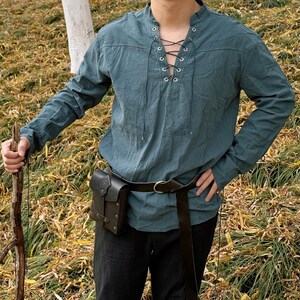 May include: A teal blue long-sleeved shirt with a lace-up neckline and a brown leather belt with a pouch. The shirt is made of a soft, comfortable fabric and has a relaxed fit.