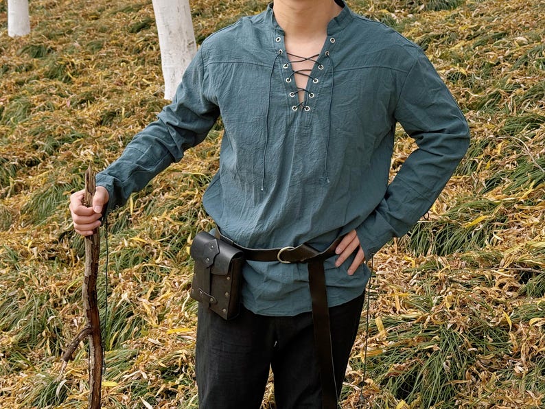 May include: A teal blue long-sleeved shirt with a lace-up neckline and a brown leather belt with a pouch. The shirt is made of a soft, comfortable fabric and has a relaxed fit.