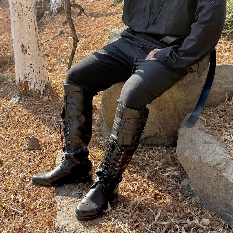 May include: A pair of black leather knee-high boots with laces and buckles. The boots are worn by a person sitting on a rock.
