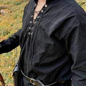 May include: A black long-sleeved shirt with a lace-up neckline and silver eyelets. The shirt has a relaxed fit and is worn with a black leather belt.