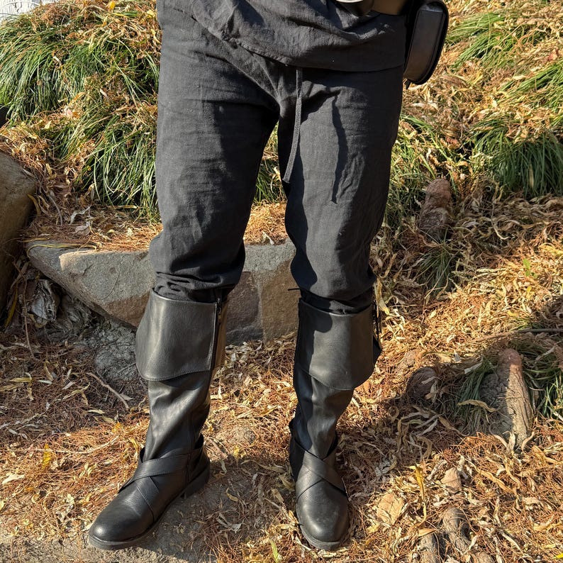 May include: A pair of black leather boots with buckles and a fold-over top. The boots are worn by a person wearing black pants.