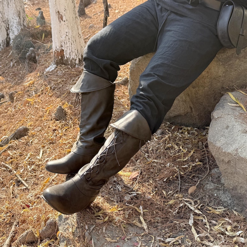 May include: A pair of brown leather boots with laces and a fold-over cuff. The boots are worn by a person wearing black pants.