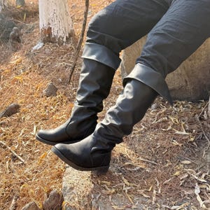 May include: A pair of black leather boots with a slouchy, folded-down design. The boots are worn by a person sitting on a rock.