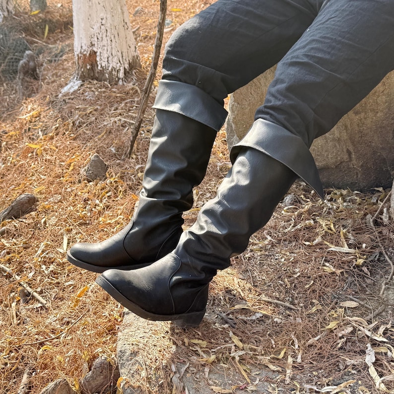 May include: A pair of black leather boots with a slouchy, folded-down design. The boots are worn by a person sitting on a rock.