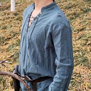 May include: A blue long-sleeved shirt with a lace-up neckline and a brown leather belt. The shirt is made of a soft, lightweight fabric and has a relaxed fit.