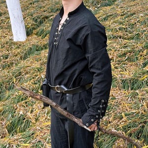 May include: A black long-sleeved shirt with a lace-up front and a leather belt. The shirt has a high collar and the sleeves are tied at the wrists. The person is holding a wooden stick.
