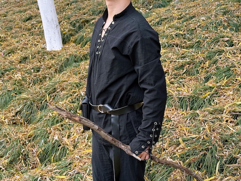 May include: A black long-sleeved shirt with a lace-up front and a leather belt. The shirt has a high collar and the sleeves are tied at the wrists. The person is holding a wooden stick.