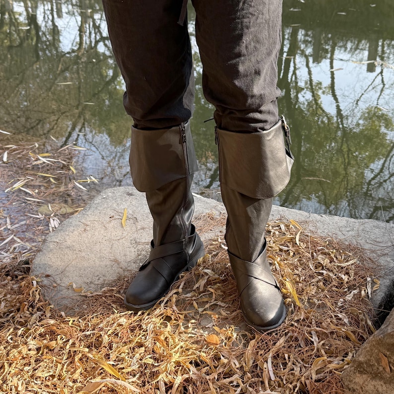 May include: Black leather boots with buckles and a fold-over cuff. The boots are worn by a person standing on a rock covered in brown leaves.