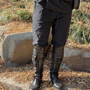 May include: Black leather knee-high boots with laces and buckles. The boots are worn by a person wearing black pants and a black belt.