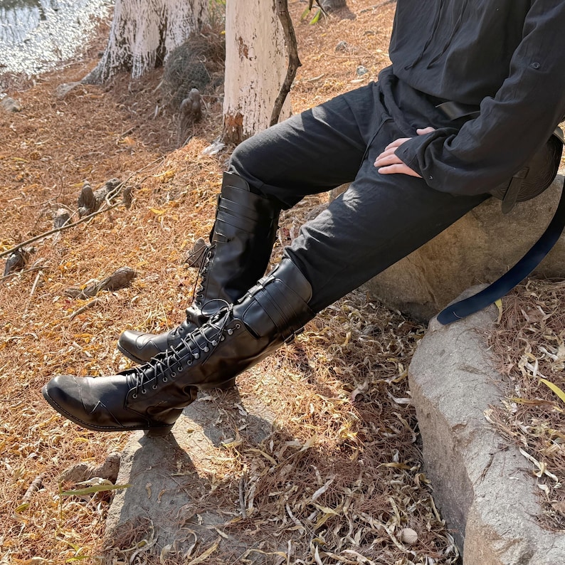 May include: Black leather boots with laces and buckles. The boots are worn by a person sitting on a rock.