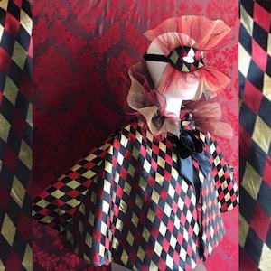 Könnte beinhalten: Ein rot-schwarz-goldenes Harlekin-Jester-Kostüm mit passender Augenmaske. Das Kostüm besteht aus einem Umhang mit Rautenmuster und einem Rüschenkragen.