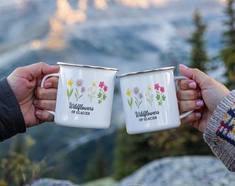 May include: Two white enamel mugs with silver rims are held up, each displaying a floral design with the text "Wildflowers of Glacier." The mugs feature a handle and are set against a blurred mountain backdrop.