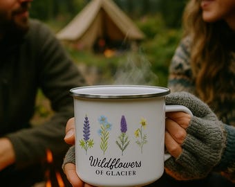 May include: White enamel camping mug with a black rim, featuring a wildflower design and the text "Wildflowers of Glacier." The mug is held near a campfire, with a tent and mountains in the background.