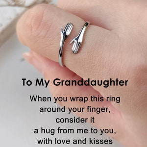 May include: A silver-colored ring with two hands embracing a finger. The ring has the text "To My Granddaughter" and a message about a hug. The ring is a sentimental gift.