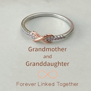 May include: A silver ring with a rose gold infinity symbol set with small clear stones. The ring is engraved with "Grandmother and Granddaughter Forever Linked Together".