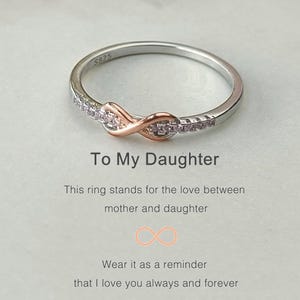 Op de afbeelding: Een zilveren ring met een roségouden oneindigheidssymbool bezet met kleine heldere stenen. De ring ligt op een witte achtergrond met de tekst "To My Daughter" en "This ring stands for the love between mother and daughter. Wear it as a reminder that I love you always and forever."