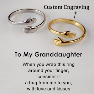 May include: Two hug rings, one silver and one gold, with the text "To My Granddaughter" and a poem about the ring being a hug from the giver. The text "Custom Engraving" is above the rings with an arrow pointing to the rings.