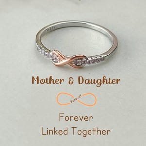 May include: A silver and rose gold infinity ring with clear cubic zirconia stones. The ring is inscribed with the words "Mother & Daughter" and "Forever Linked Together".