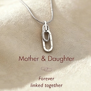 Op de afbeelding: Een zilveren ketting met twee in elkaar grijpende ovale hangers. Een van de hangers is bezet met kleine, heldere stenen. De woorden "Mother & Daughter" en "Forever linked together" staan eronder.