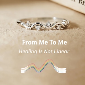 Op de afbeelding: Een zilveren ring met een golvend ontwerp, bezet met meerdere kleine, heldere edelstenen. De ring wordt gepresenteerd tegen een neutrale achtergrond, vergezeld van de tekst "From Me To Me" en "Healing Is Not Linear". Een regenbooggolfafbeelding is ook zichtbaar.