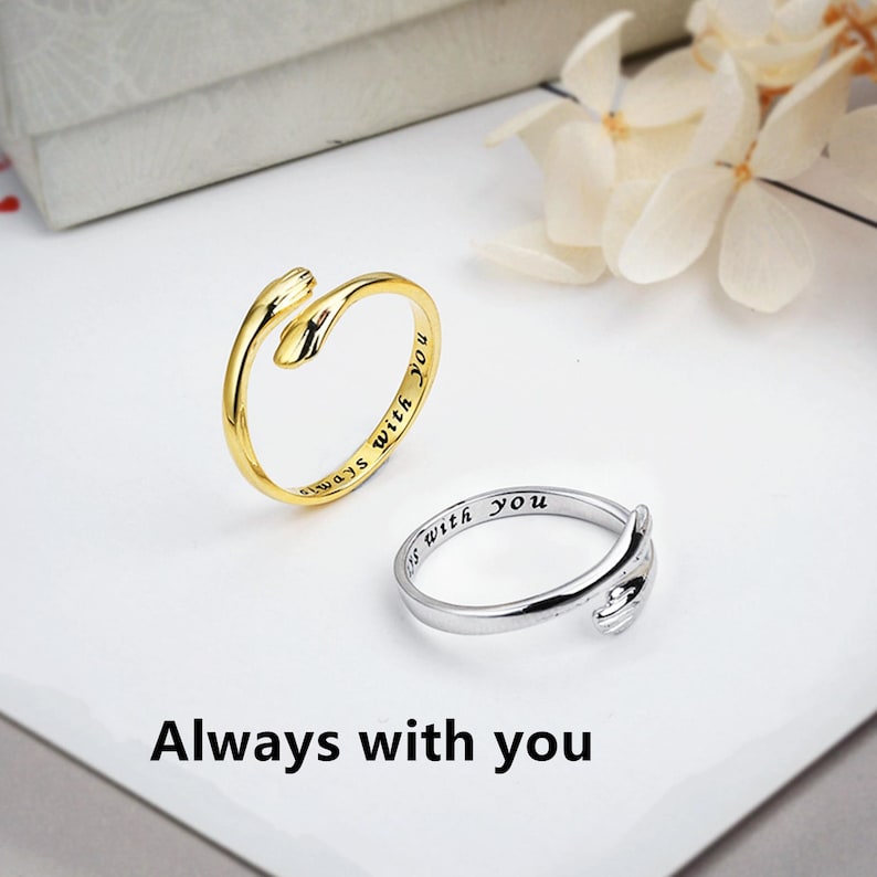 Puede incluir: Dos anillos abiertos, uno de tono dorado y otro de tono plateado, con un dise&ntilde;o de manos que se abrazan. Las bandas exteriores est&aacute;n grabadas con el texto Always with you en una fuente cursiva negra. Los anillos tienen un acabado pulido.