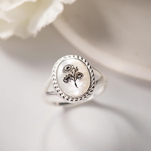 K&ouml;nnte beinhalten: Silberring mit ovaler Perlmutt-Oberfl&auml;che. Die Oberfl&auml;che zeigt ein schwarzes Blumendesign. Der Ring hat einen geflochtenen Rand und einen geteilten Schaft. Der Ring ist vor einem wei&szlig;en Hintergrund platziert.