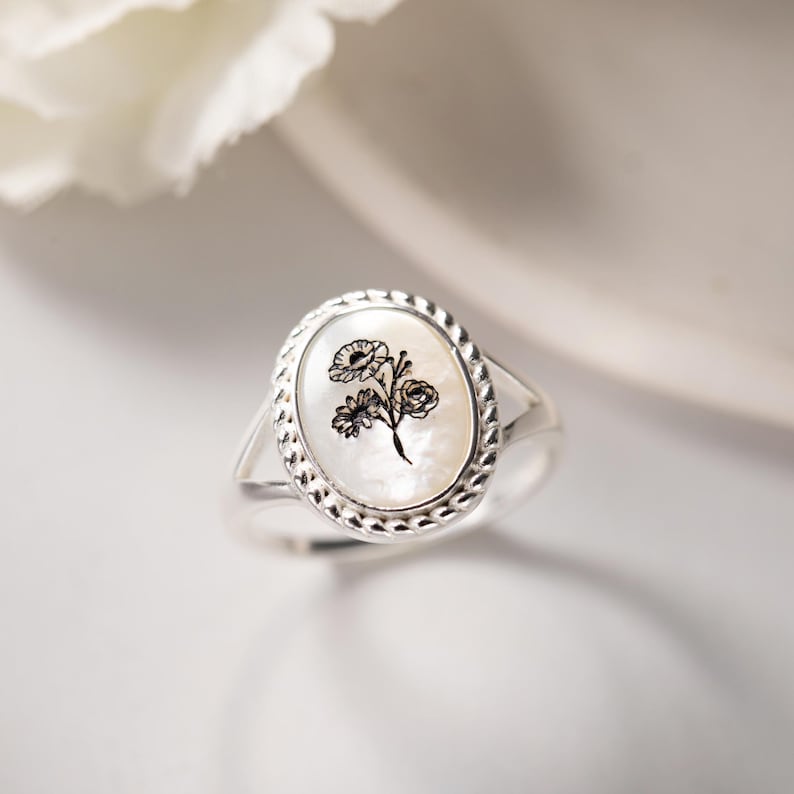 K&ouml;nnte beinhalten: Silberring mit ovaler Perlmutt-Oberfl&auml;che. Die Oberfl&auml;che zeigt ein schwarzes Blumendesign. Der Ring hat einen geflochtenen Rand und einen geteilten Schaft. Der Ring ist vor einem wei&szlig;en Hintergrund platziert.