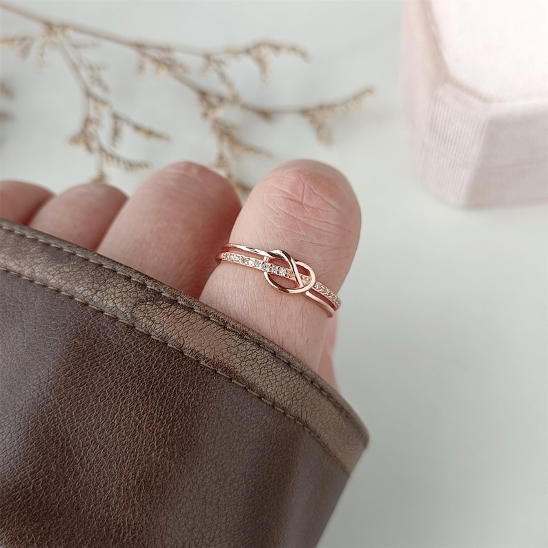 May include: A rose gold ring with a knot design and a row of small clear stones.