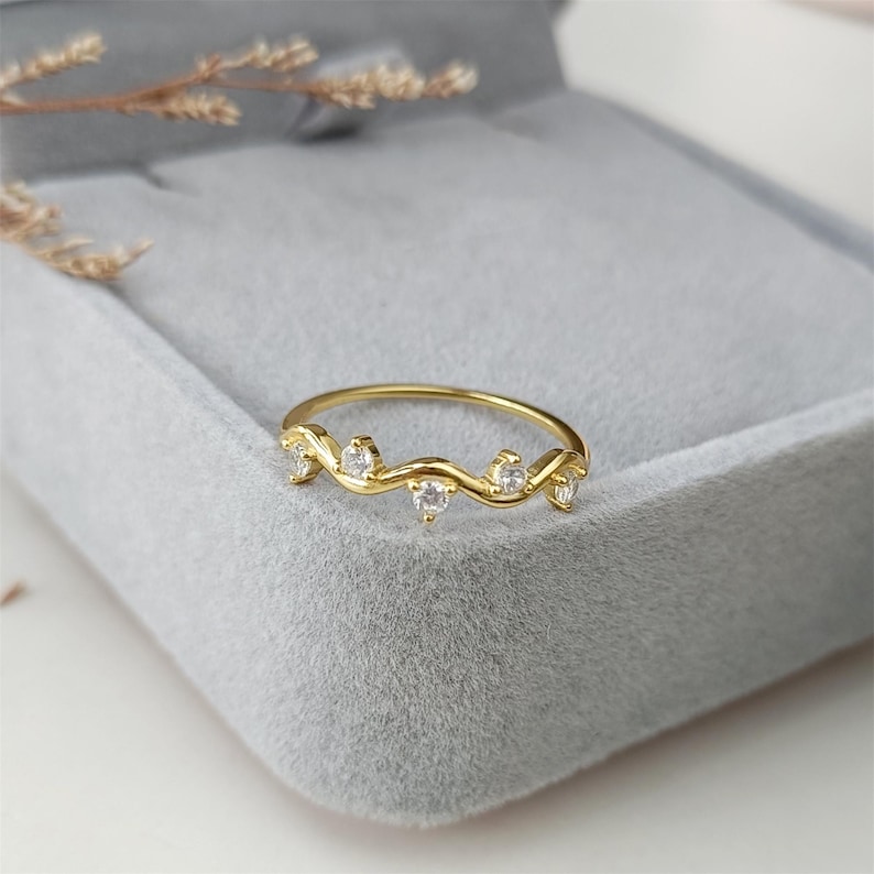 Puede incluir: Un delicado anillo de oro con una banda ondulada y cinco peque&ntilde;as piedras transparentes.