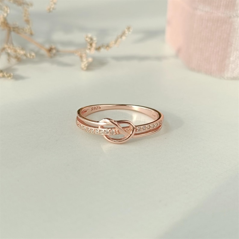 May include: A rose gold ring with a knot design and small clear stones. The ring is marked with the number "S925".