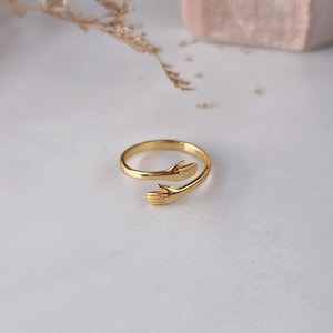 Puede incluir: Un anillo de tono dorado con un dise&ntilde;o de banda abierta donde los dos extremos tienen forma de peque&ntilde;as manos que se extienden una hacia la otra. El metal tiene un acabado liso y pulido, y el anillo es una pieza de metal continua.
