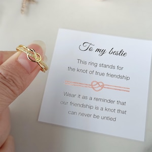 May include: Gold ring with a knot design and small clear stones. The ring is on a finger and next to a card that says "To my bestie" and "This ring stands for the knot of true friendship. Wear it as a reminder that our friendship is a knot that can never be untied."