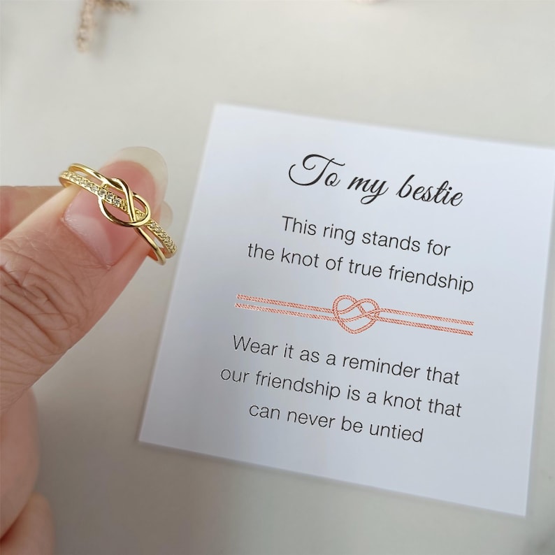 May include: Gold ring with a knot design and small clear stones. The ring is on a finger and next to a card that says "To my bestie" and "This ring stands for the knot of true friendship. Wear it as a reminder that our friendship is a knot that can never be untied."