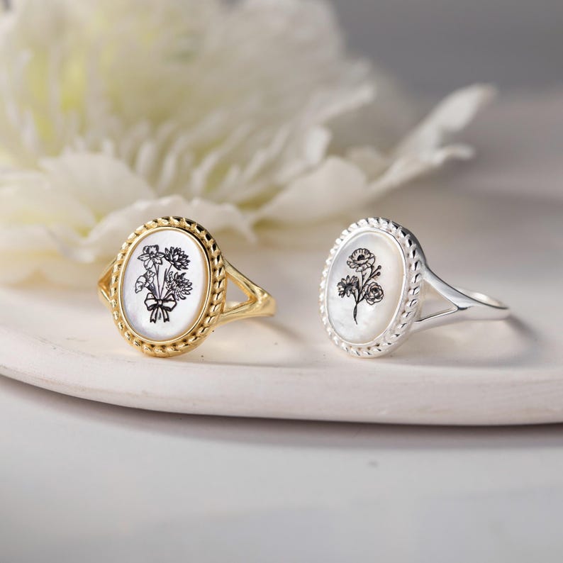 K&ouml;nnte beinhalten: Zwei ovale Ringe mit floralen Designs. Ein Ring ist goldfarben mit einer Blumenstrau&szlig;-Illustration, der andere silberfarben mit einer einzelnen Blume. Beide Ringe haben einen wei&szlig;en Hintergrund und einen seilartigen Rand.
