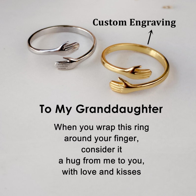 Puede incluir: Dos anillos abiertos, uno plateado y otro dorado, cada uno con dos manos que se encuentran en un dise&ntilde;o de abrazo. El texto dice: Custom Engraving, To My Granddaughter, When you wrap this ring around your finger, consider it a hug from me to you, with love and kisses.