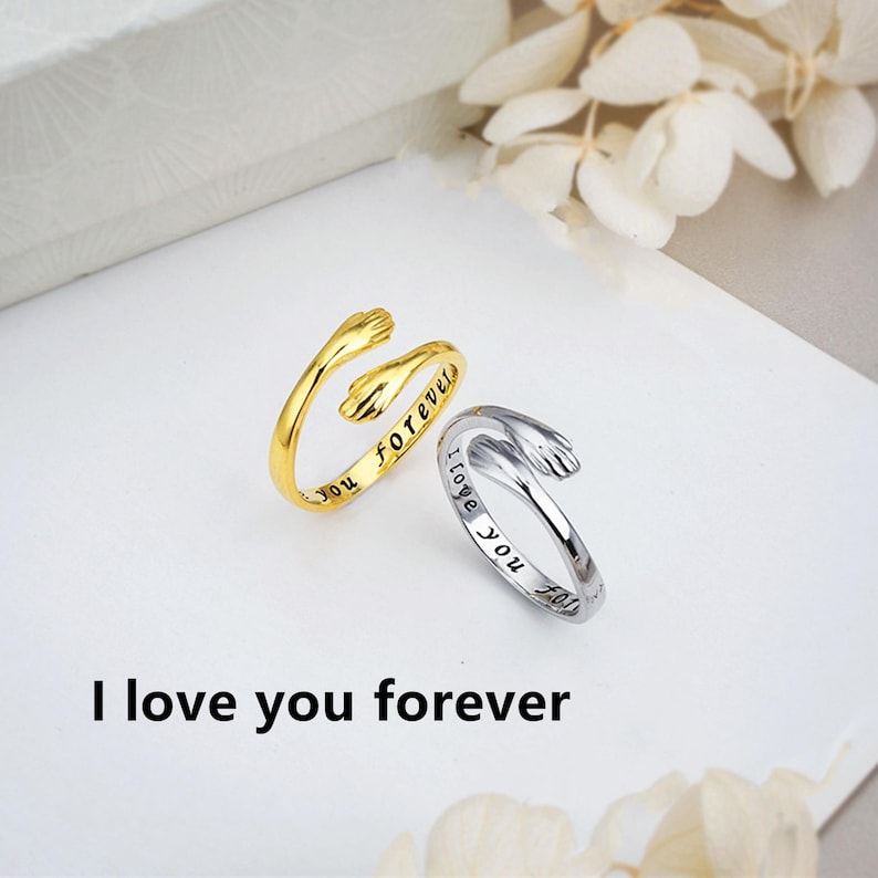 Puede incluir: Dos anillos abiertos con un dise&ntilde;o de manos que se abrazan. Un anillo es de tono dorado con el texto grabado 'love you forever' en la banda interior, y el otro es de tono plateado con el texto grabado 'I love you' en la banda interior.